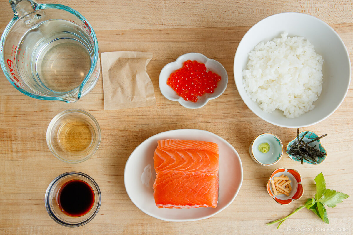 Top-down view of ingredients for a Japanese dish: raw salmon, cooked white rice, salmon roe, soy sauce, mirin, water, seasoning packet, sliced ginger, wasabi, and pieces of dried seaweed on a wooden surface.