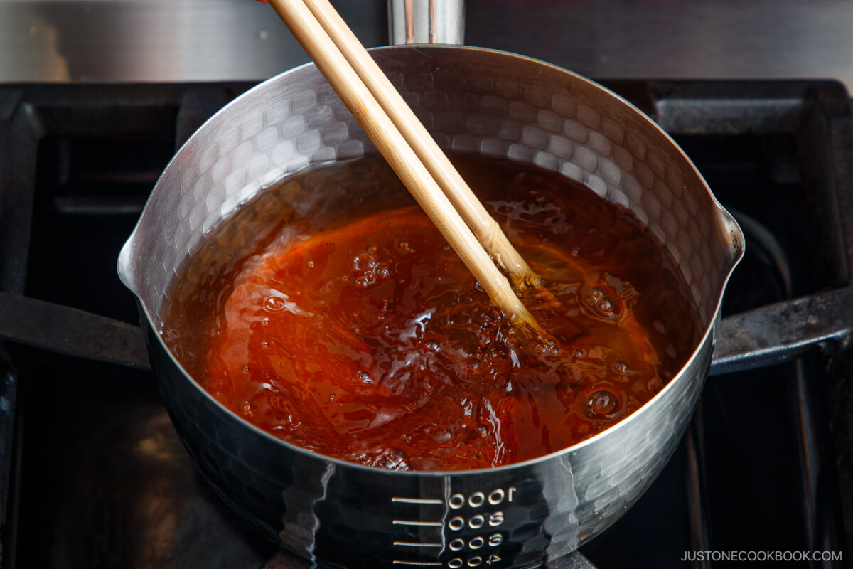 A metal saucepan filled with simmering brown liquid is being stirred with a pair of wooden chopsticks on a stovetop.