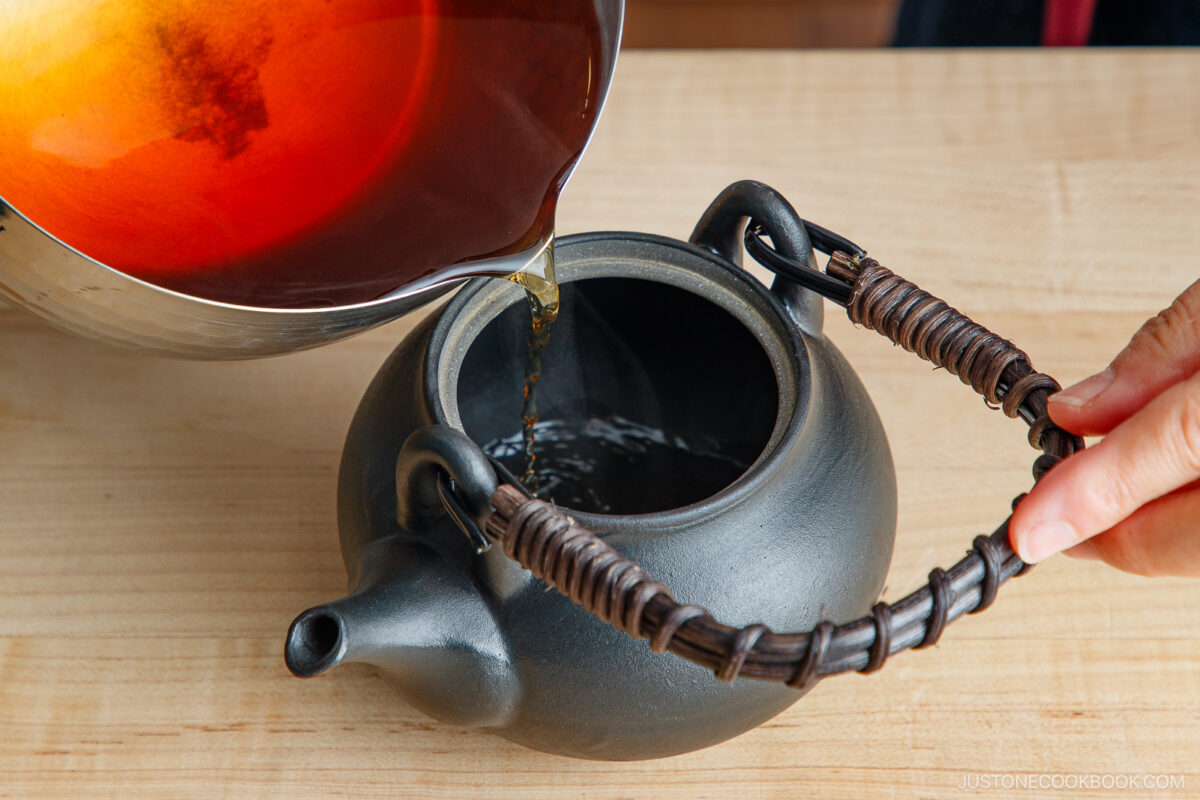 A person pours hot tea from a metal bowl into a black teapot with a wrapped handle, all set on a wooden surface.