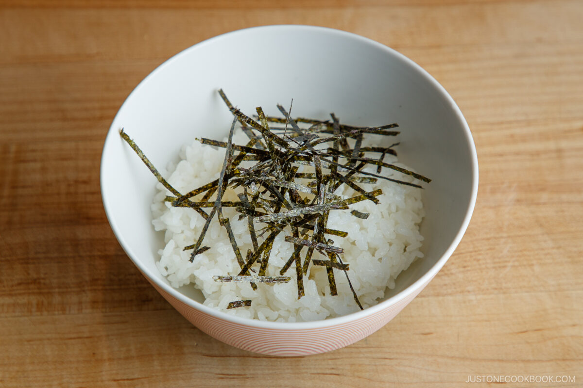 A white bowl of steamed white rice topped with thinly sliced strips of nori seaweed, placed on a wooden surface.