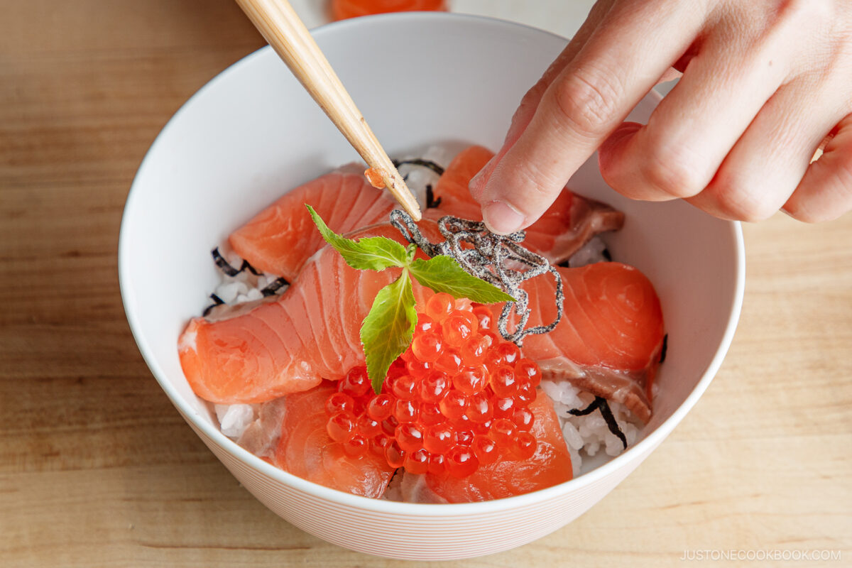 A hand uses chopsticks to add garnish to a bowl of rice topped with salmon sashimi, salmon roe, and a green leaf on a wooden surface.