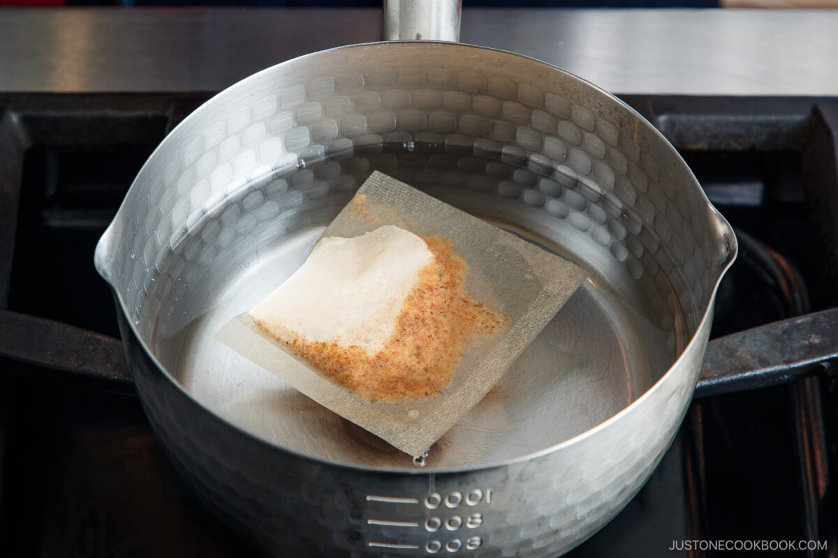 A packet of dashi soup base is simmering in a stainless steel pot filled with water on a stovetop.