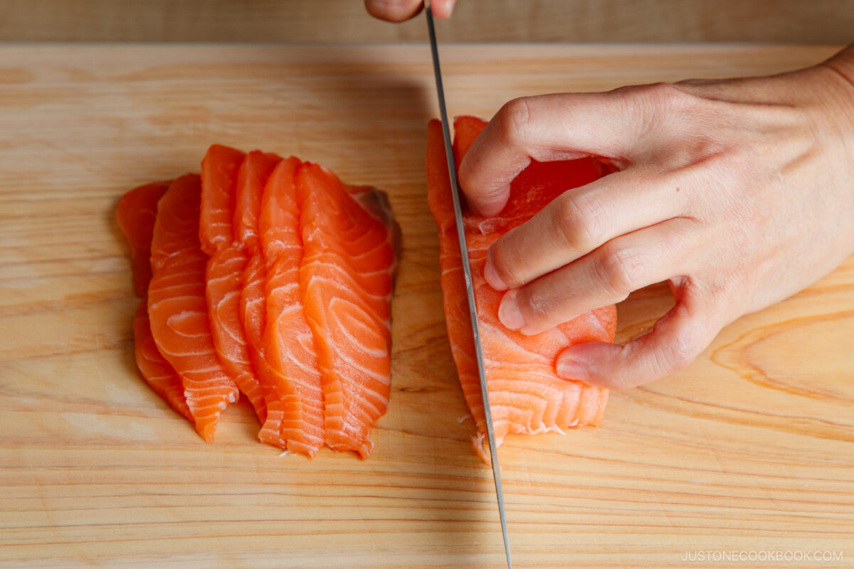A person’s hand slices raw salmon fillets into thin pieces on a wooden cutting board, with already sliced pieces neatly arranged beside the knife.