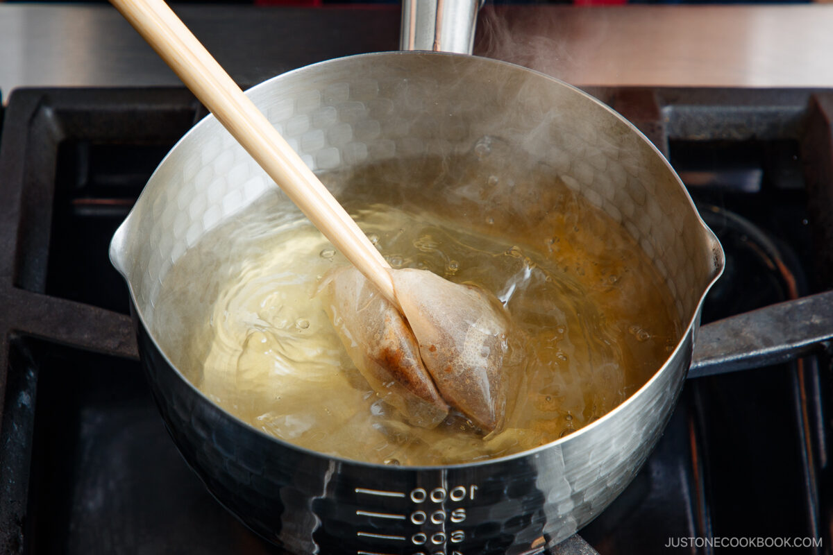 A wooden spoon stirs a teabag in a pot of hot water on a stovetop, with steam rising from the pot.