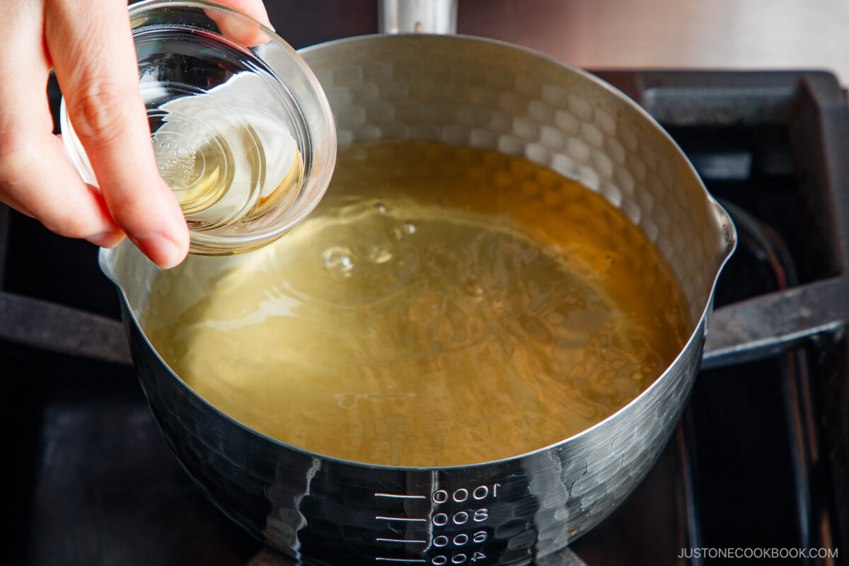 A hand pours liquid from a small glass bowl into a pot of clear broth heating on a stovetop.