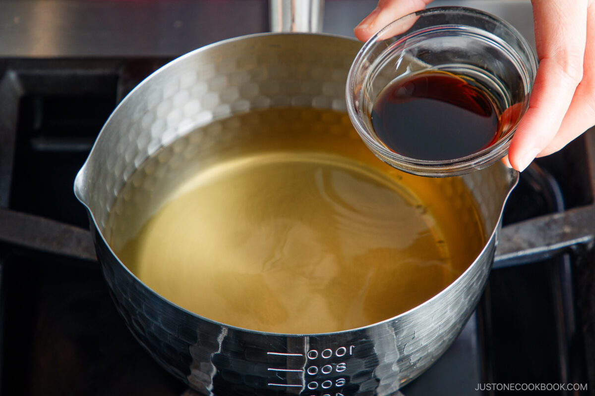 A hand holds a small glass bowl of dark liquid above a saucepan filled with clear broth on a stovetop, preparing to pour the liquid into the pot.