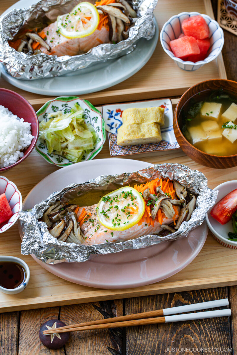 A Japanese meal with salmon and vegetables baked in foil, miso soup, steamed rice, pickled cabbage, rolled omelette, and watermelon pieces, all served on a wooden tray with chopsticks and a dipping sauce.