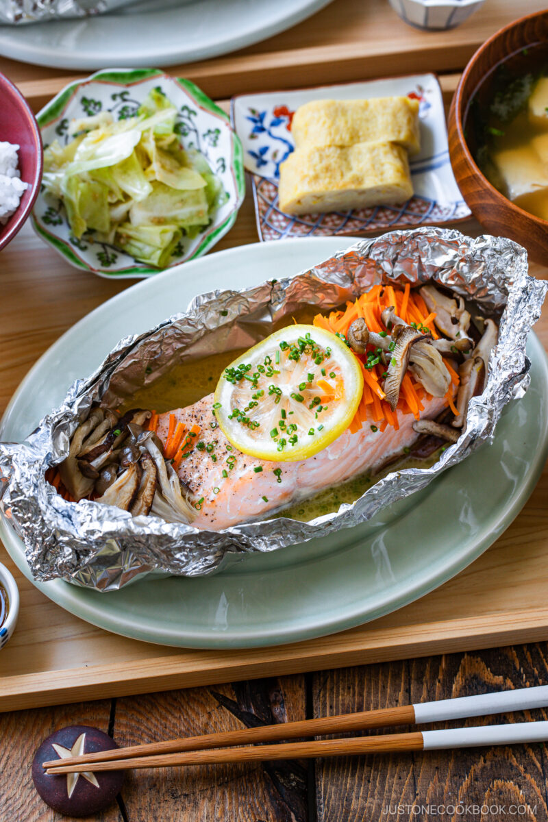 Baked salmon fillet with lemon, mushrooms, and carrots served in foil on a plate, accompanied by sides of lettuce salad, tamagoyaki, miso soup, and white rice on a wooden table.