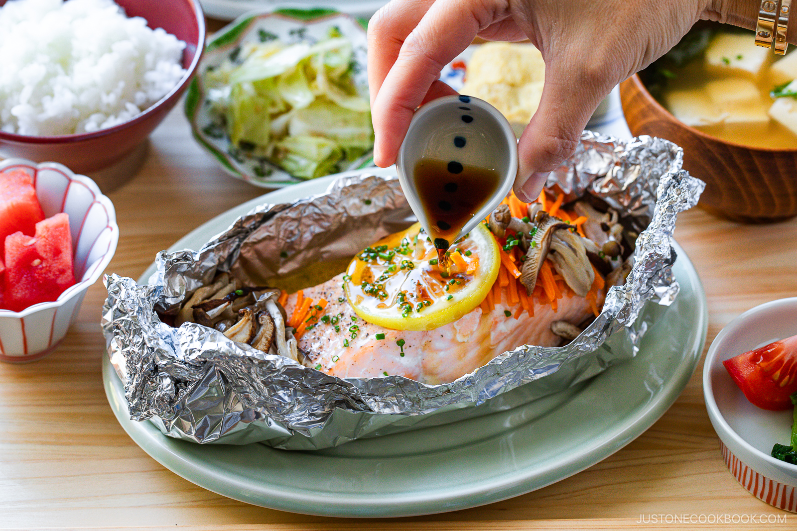 A hand pours sauce over baked salmon with lemon, mushrooms, and veggies in foil. Surrounding dishes include rice, salad, soup, watermelon, and tomato on a wooden table.