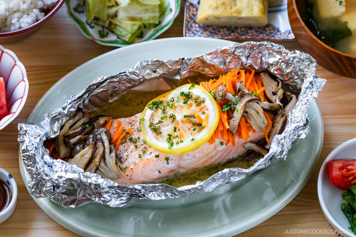 Baked salmon fillet topped with lemon slices, shredded carrots, and mushrooms, served in aluminum foil on a plate, surrounded by various side dishes on a wooden table.