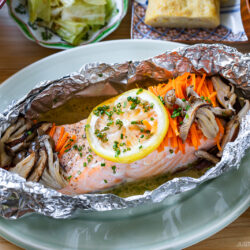 Baked salmon fillet topped with lemon slices, shredded carrots, and mushrooms, served in aluminum foil on a plate, surrounded by various side dishes on a wooden table.