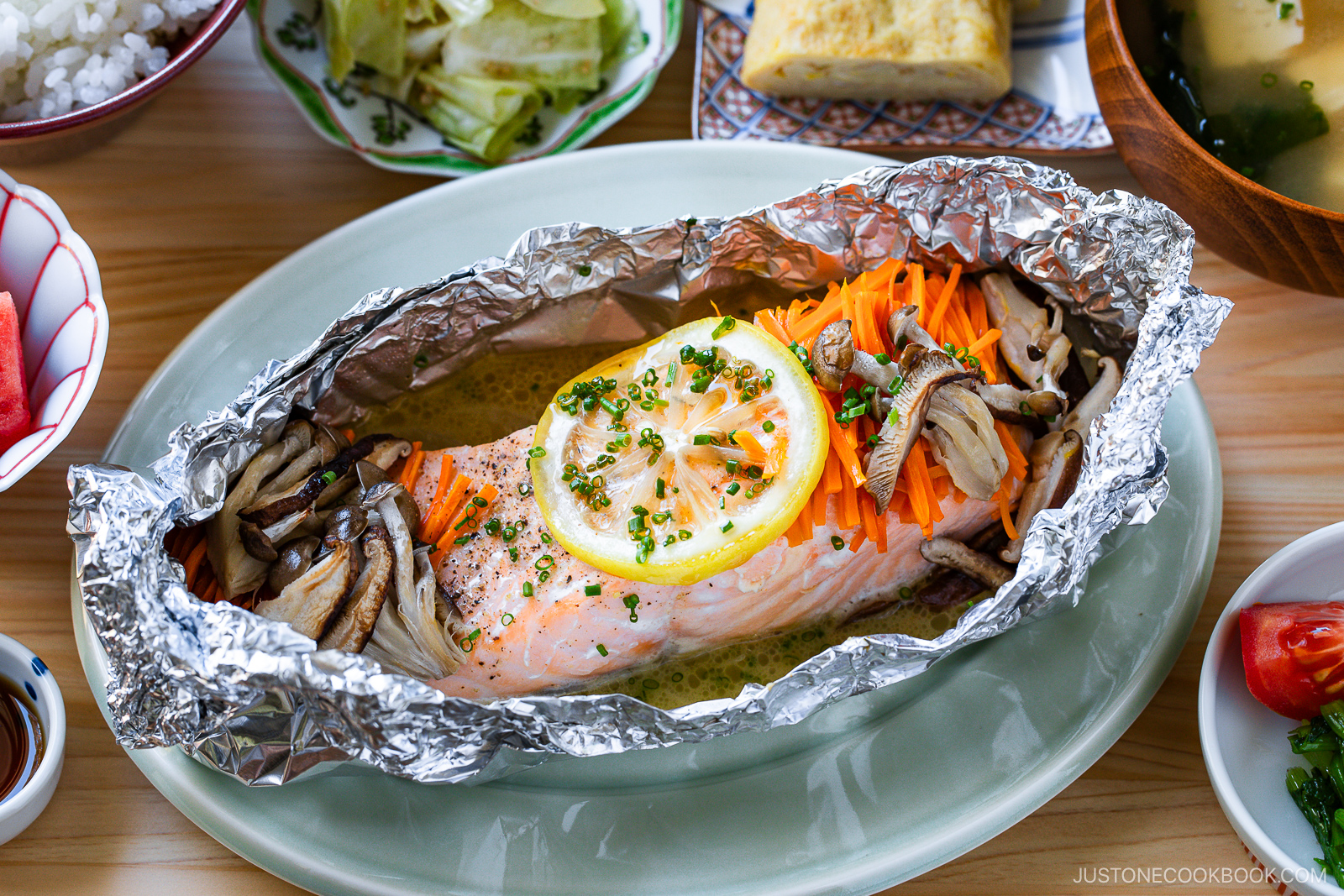 Baked salmon fillet topped with lemon slices, shredded carrots, and mushrooms, served in aluminum foil on a plate, surrounded by various side dishes on a wooden table.