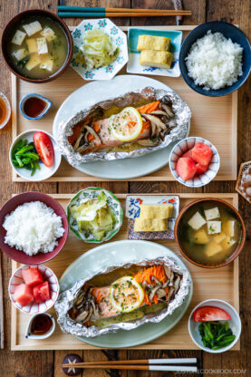 A Japanese meal set with two trays, each featuring foil-baked salmon with vegetables, rice, miso soup, tamagoyaki, pickles, sliced tomato, watermelon, and various small side dishes, all arranged neatly on a wooden table.