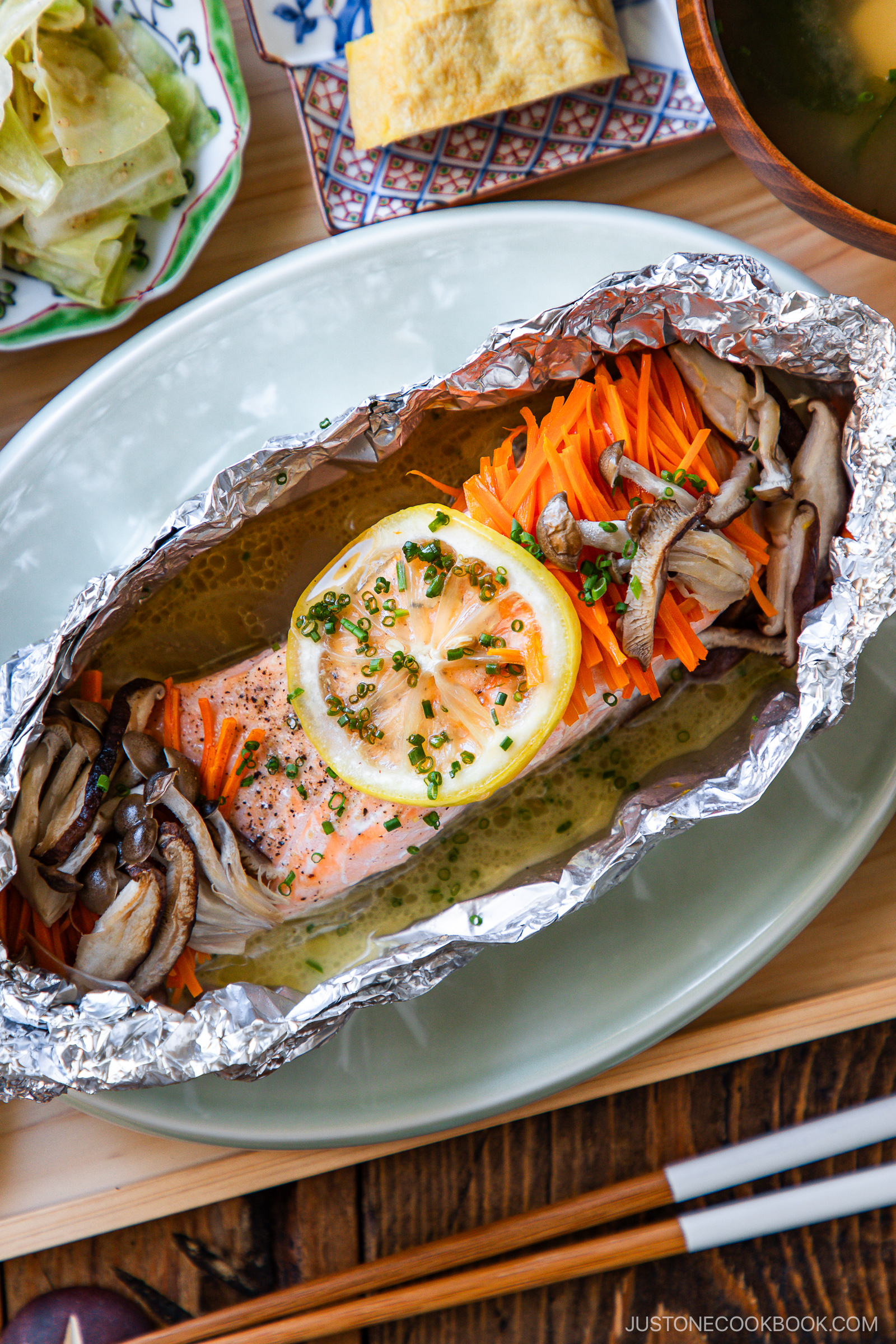 Salmon fillet baked in foil with sliced lemon, julienned carrots, and mushrooms, garnished with chives. Japanese side dishes, rice, and chopsticks are visible in the background.