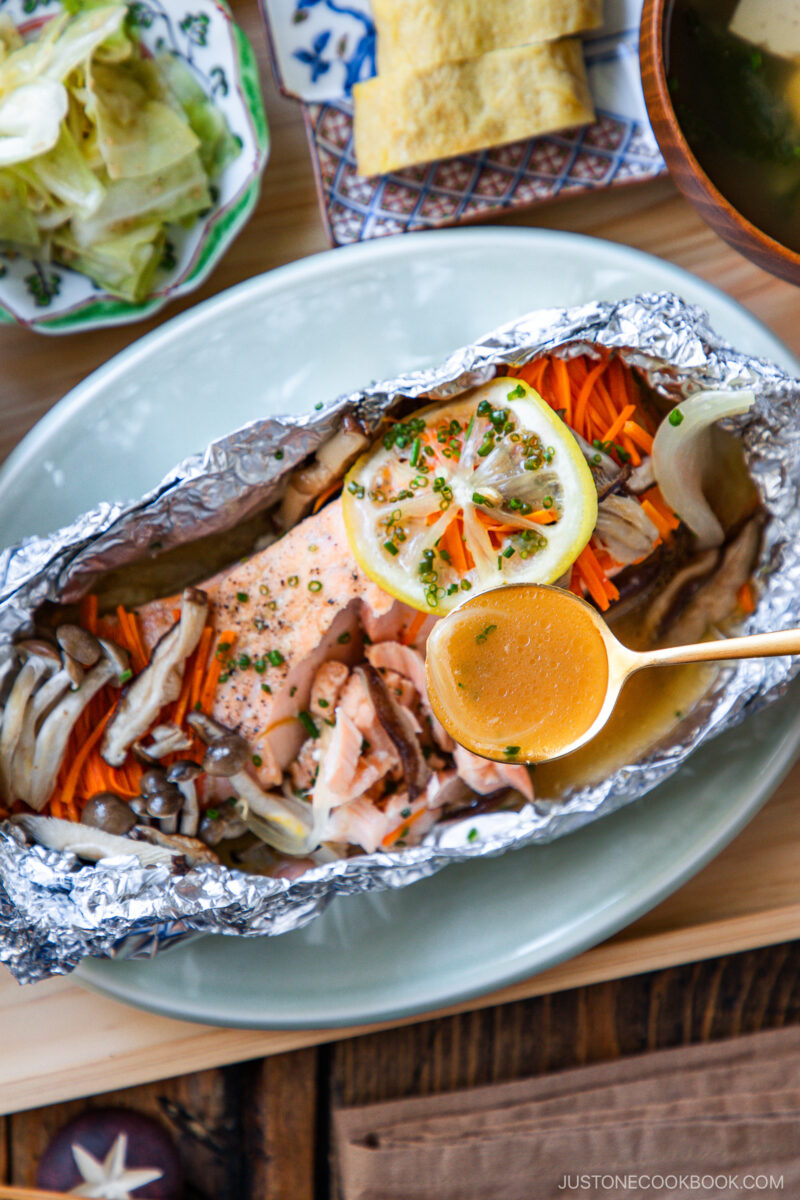 Salmon fillet baked in foil with mushrooms, carrots, onions, and lemon slices, garnished with chives, with a spoon pouring sauce over it. The dish is served on a plate with salad and tofu sides nearby.