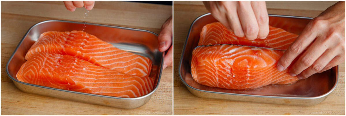 Two side-by-side images showing hands seasoning raw salmon fillets in a metal tray with salt on a wooden surface. The left image shows sprinkling salt; the right image shows rubbing salt into the fish.
