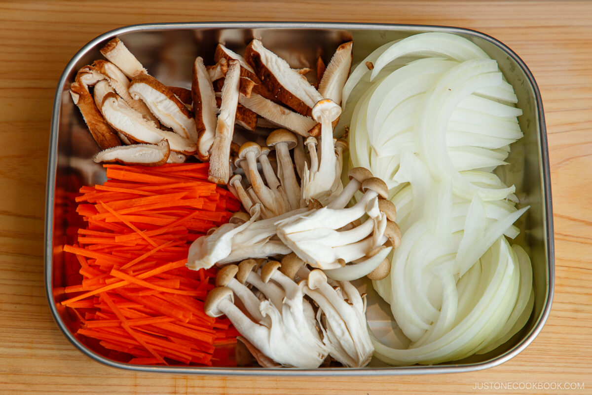 A metal tray with neatly arranged sliced shiitake mushrooms, julienned carrots, shimeji mushrooms, and thinly sliced onions on a wooden surface.