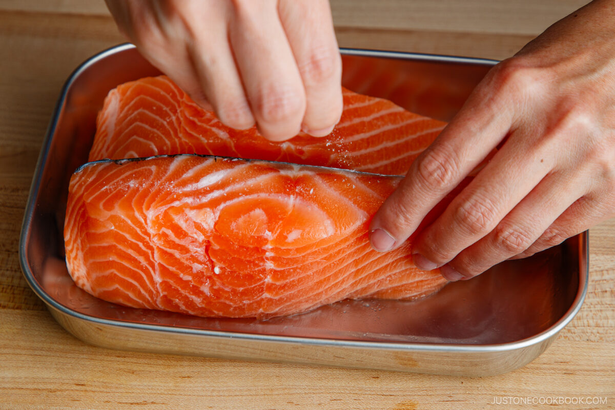 A person sprinkles salt onto two raw salmon fillets placed in a metal tray on a wooden surface.