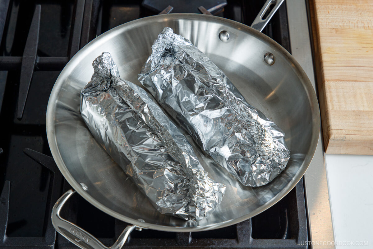 Two ears of corn wrapped in aluminum foil sit in a stainless steel pan on a stovetop, ready to be cooked. A wooden cutting board is visible to the side.