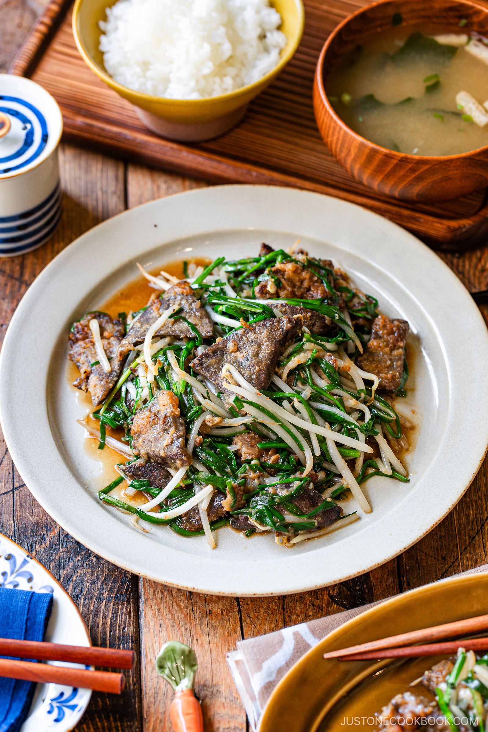 A plate of stir-fried beef with bean sprouts and green vegetables is served on a table with a bowl of white rice, a bowl of soup, and various side dishes. Chopsticks and a napkin are placed nearby.