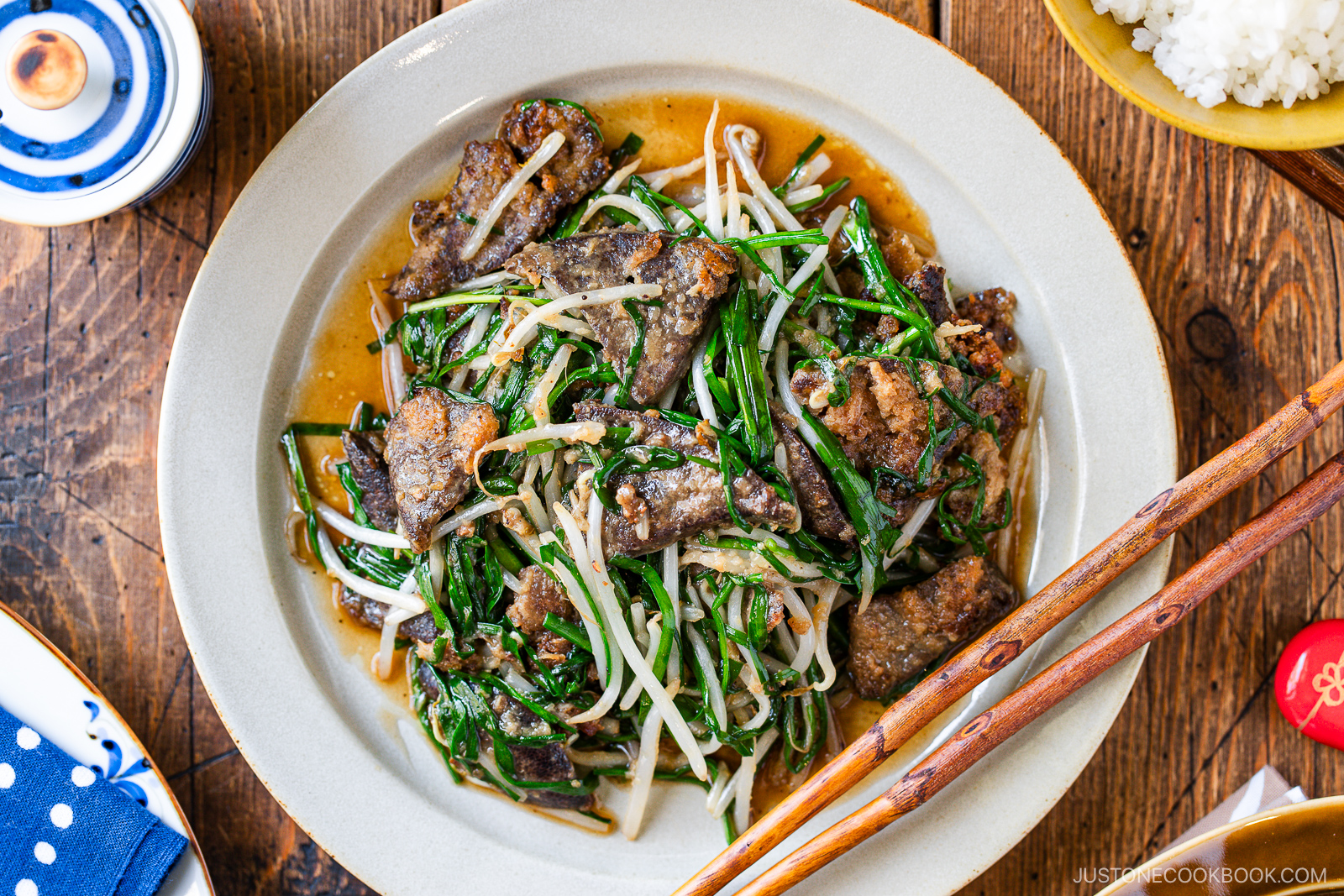 A plate of stir-fried beef liver with bean sprouts and green vegetables, served with chopsticks on a wooden table. A bowl of rice and small dish of pickles are nearby.