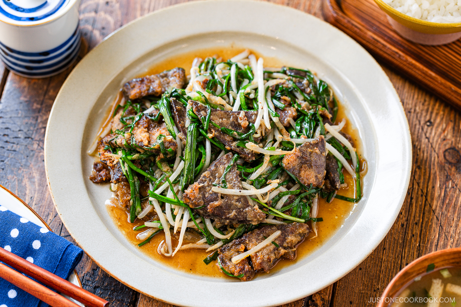 A plate of stir-fried beef liver with Chinese chives and bean sprouts in a savory sauce, served on a beige dish on a wooden table, with bowls and chopsticks nearby.