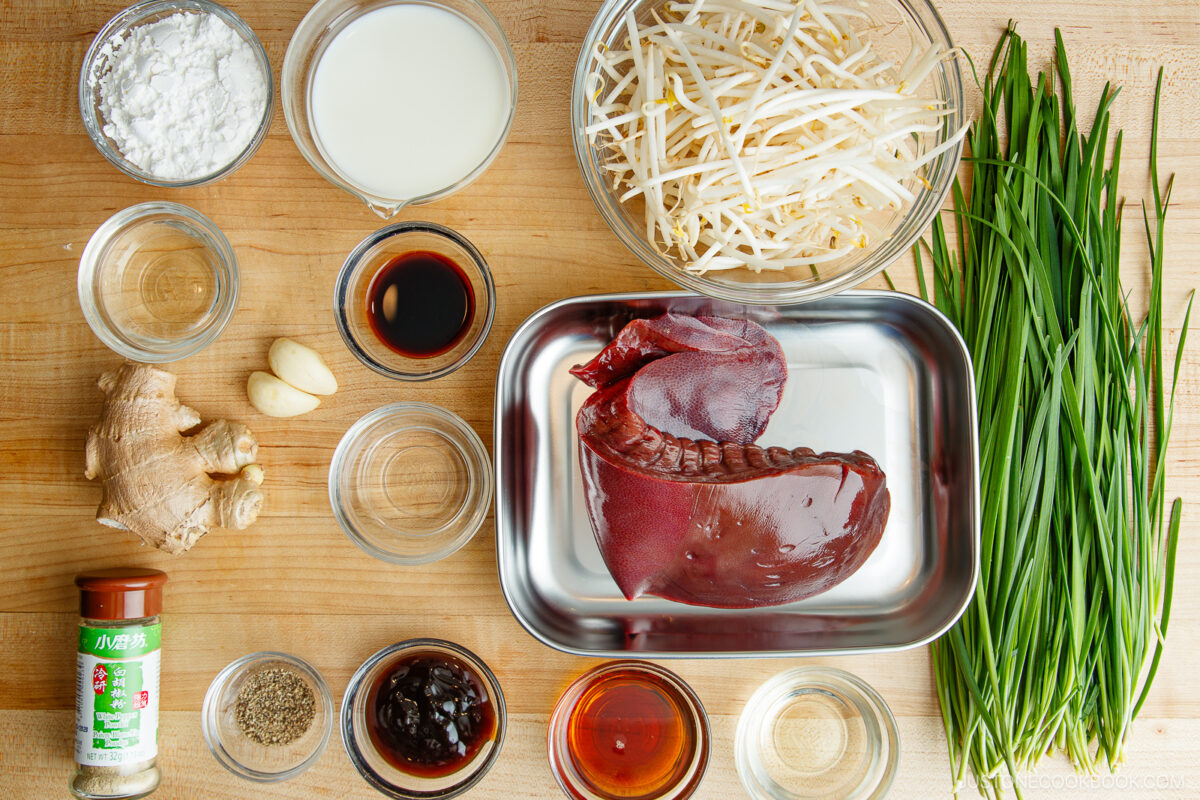 A top-down view of neatly arranged ingredients on a wooden surface: raw pork liver, fresh garlic chives, bean sprouts, ginger, garlic, various sauces, spices, cornstarch, milk, and small bowls with liquids.