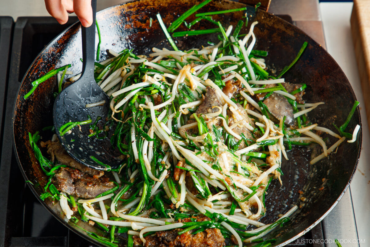A hand stirs a wok filled with stir-fried beef, bean sprouts, and green vegetables on a stove, using a black spatula.