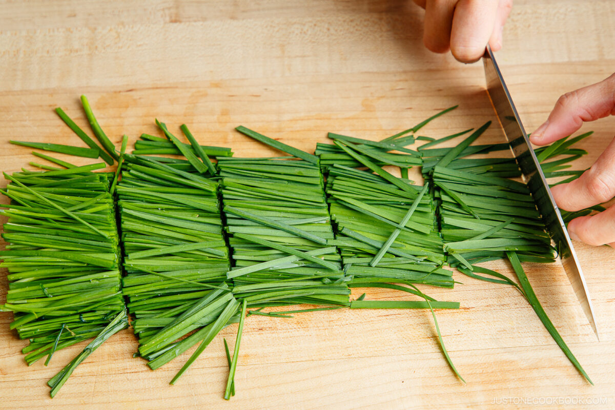 A person is using a knife to slice a bundle of green chives on a wooden cutting board.