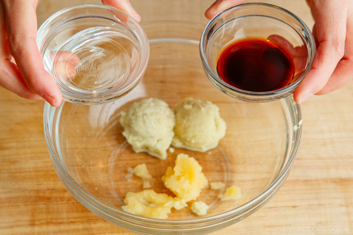 A person holding a small bowl of clear liquid and a bowl of dark liquid over a glass mixing bowl containing scoops of grated ginger and garlic on a wooden surface.