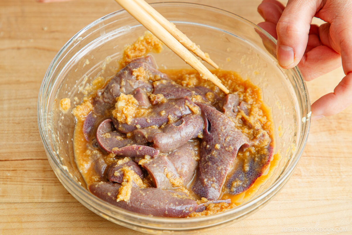 A hand uses chopsticks to mix slices of raw liver in a glass bowl with a marinade of grated ginger and seasoning on a wooden surface.