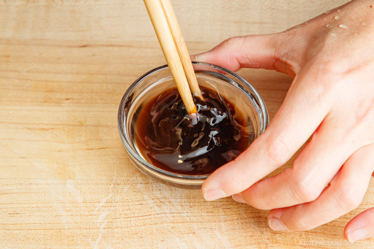 A hand stirs a thick, dark brown sauce in a small glass bowl using chopsticks, on a light wooden surface.