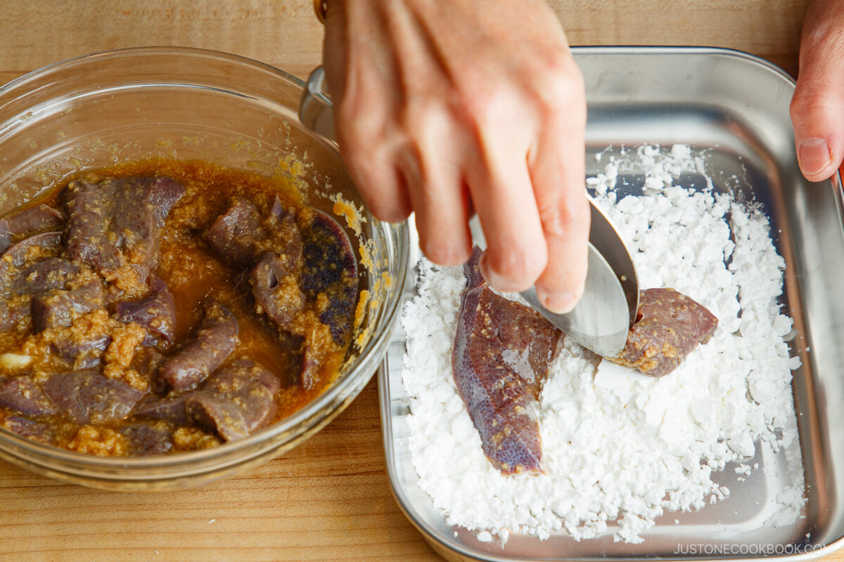 A hand coats a marinated meat slice in white starch on a metal tray, with more marinated meat in a glass bowl nearby, on a wooden surface.