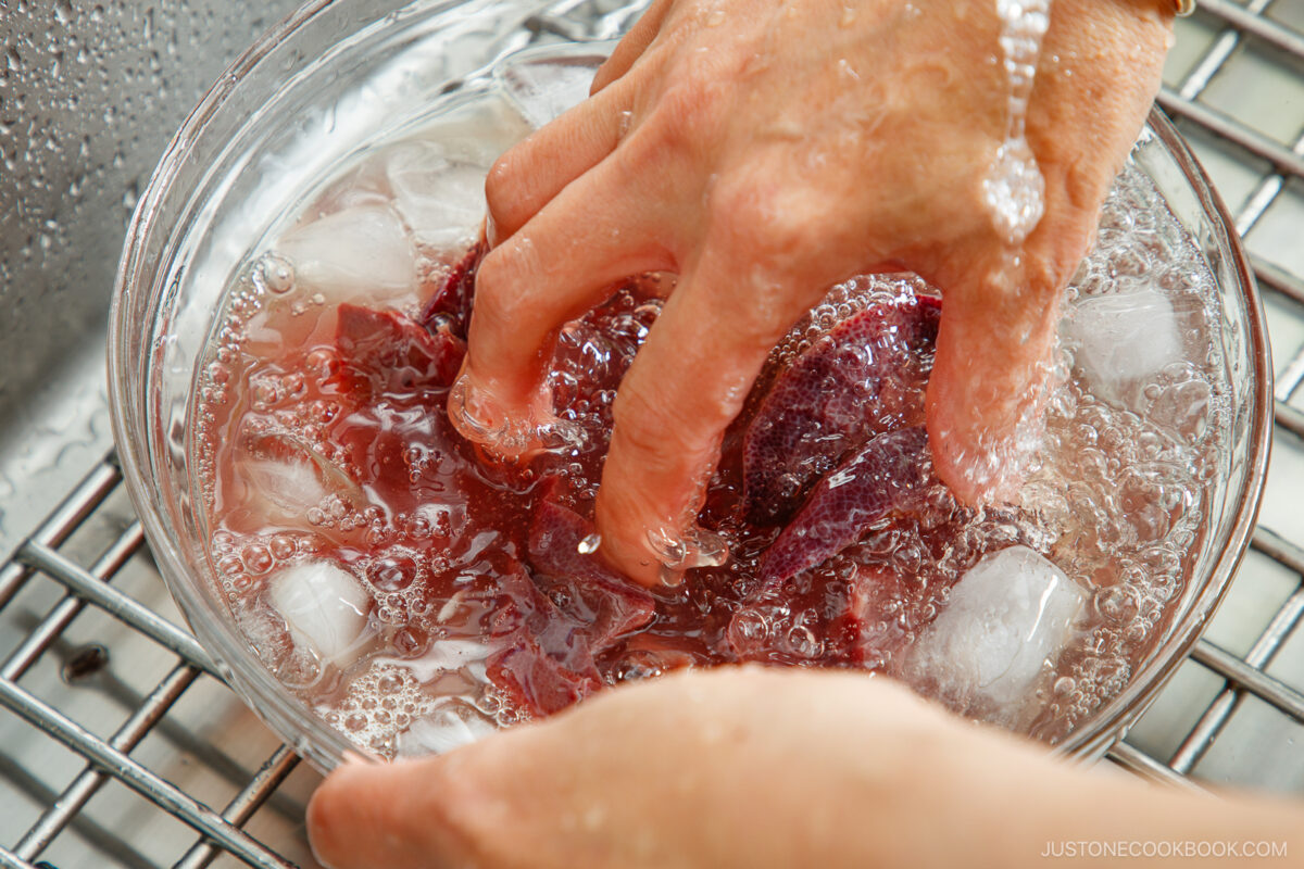 Hands submerging red beet pieces in a bowl of ice water, with ice cubes floating and water splashing, on a metal rack.
