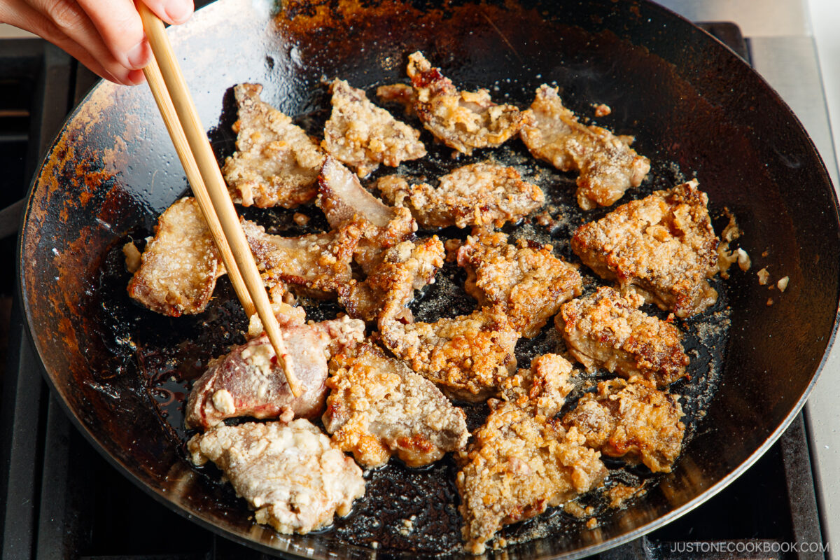 A hand uses chopsticks to cook and turn pieces of breaded meat sizzling in a large, black frying pan on a stovetop.