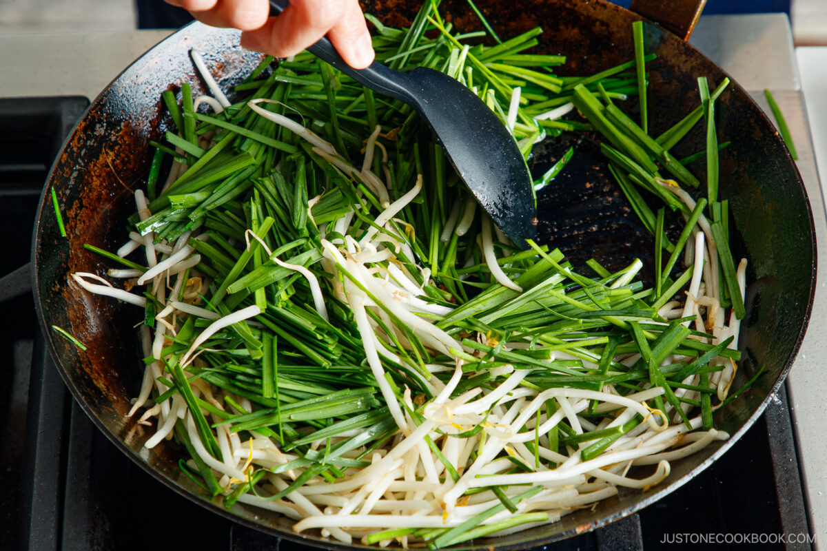 A close-up of a frying pan on a stove with bean sprouts and chives being stirred by a person using a black spatula.