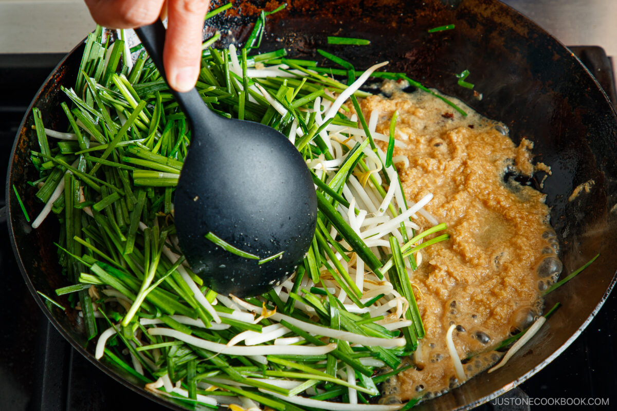 A hand holds a black ladle over a skillet with green vegetables, bean sprouts, and a bubbling golden batter cooking on the stovetop.