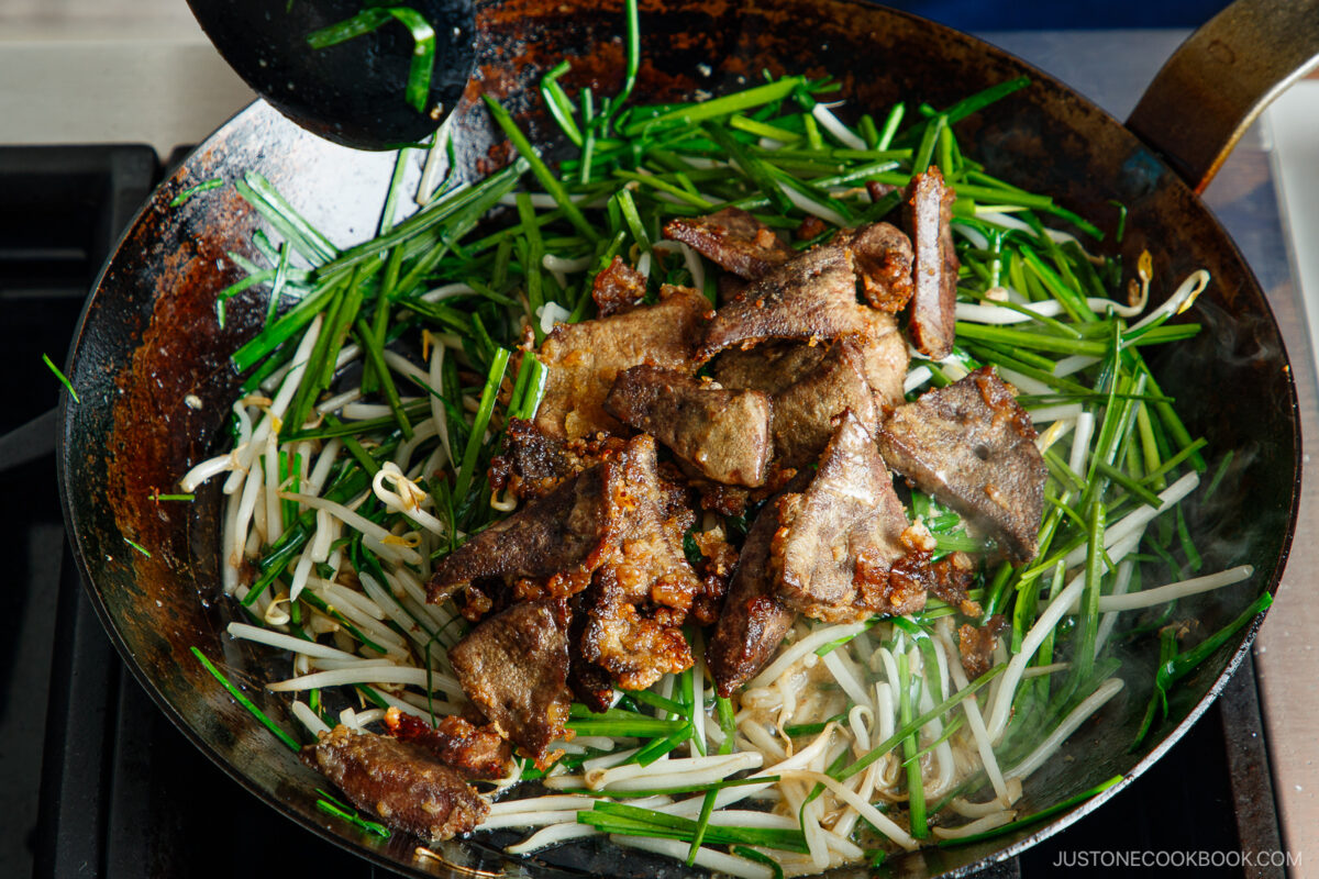 A wok on a stove filled with sautéed beef liver slices, bean sprouts, and green garlic chives, being cooked together.