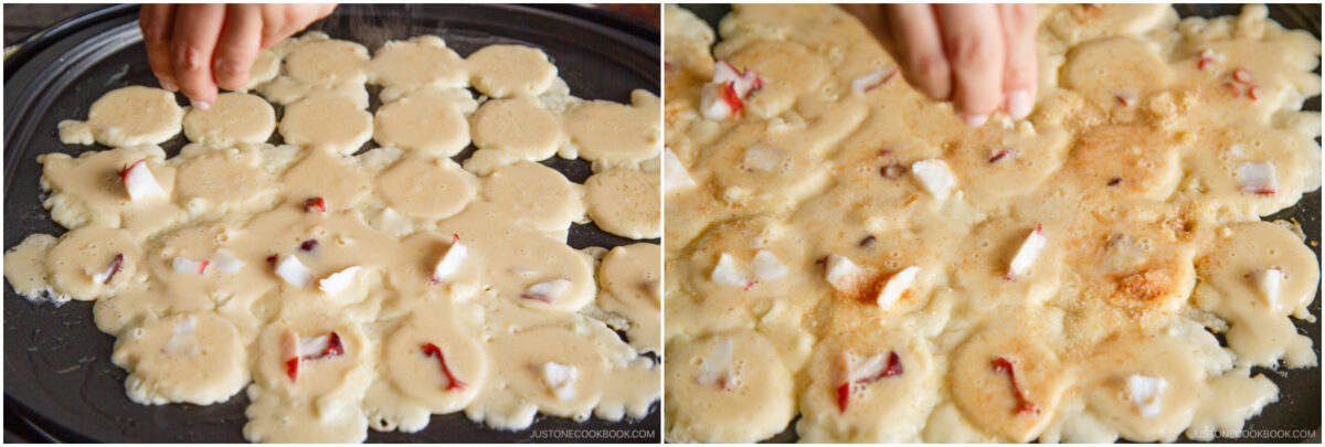 Close-up of a hand sprinkling seasoning over small, round batter portions with pieces of seafood, likely cooking takoyaki or a similar dish, on a griddle.