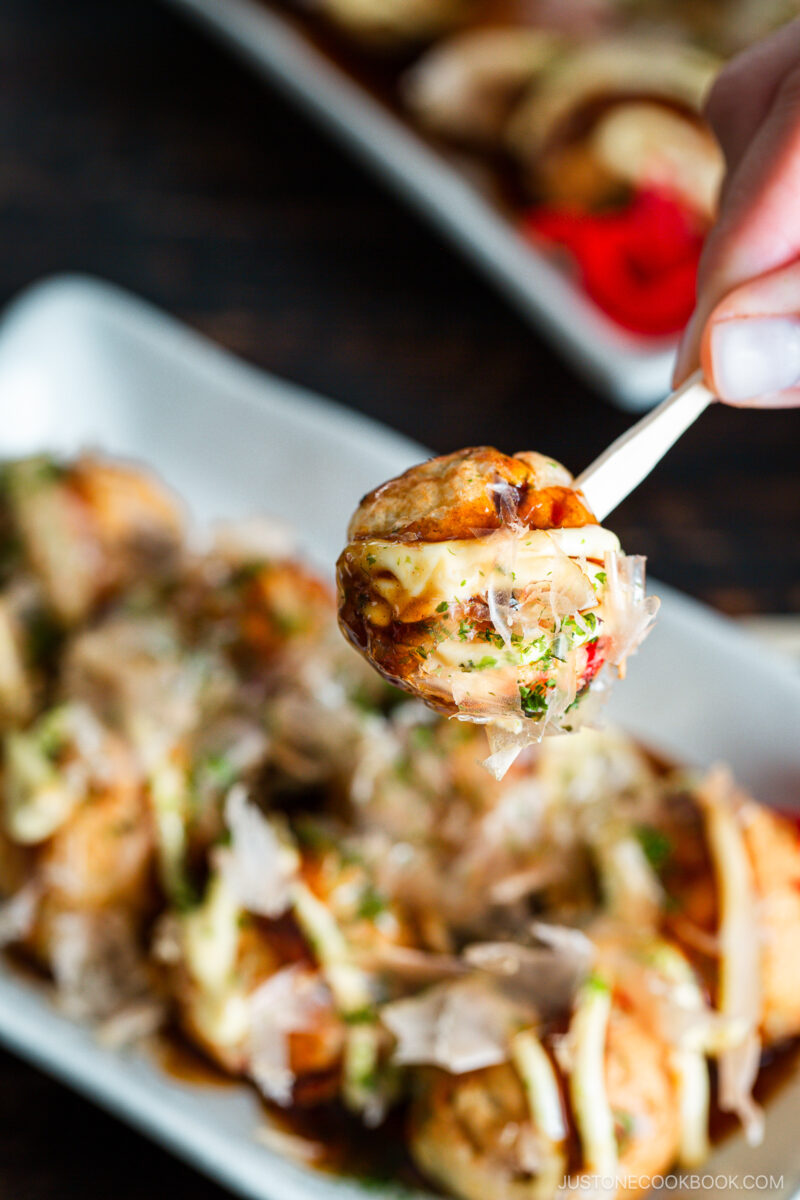 A close-up of a hand holding a toothpick with a takoyaki ball drizzled with mayonnaise and sauce, with more takoyaki balls garnished with bonito flakes on a white plate in the background.