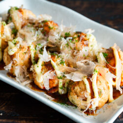 A rectangular plate of takoyaki, Japanese octopus balls, topped with mayonnaise, takoyaki sauce, bonito flakes, seaweed, and garnished with red pickled ginger on the side.