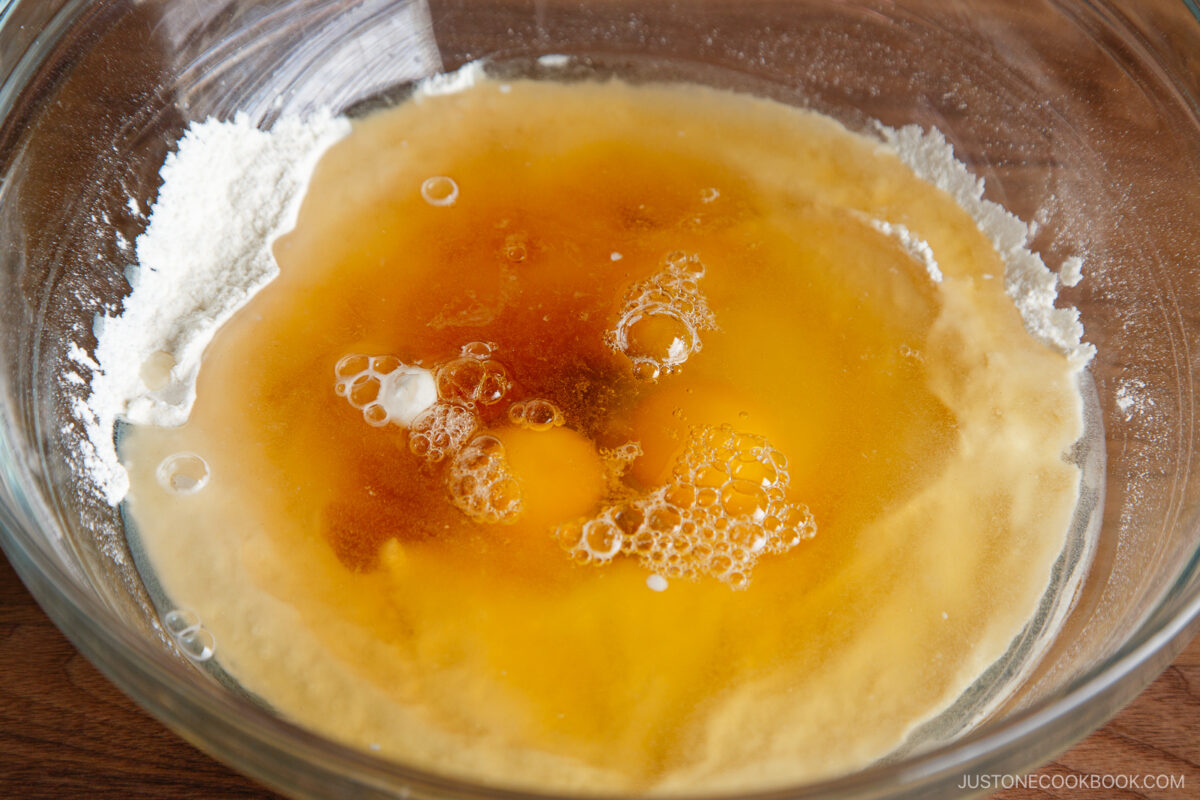 A glass mixing bowl containing flour, eggs, and liquid ingredients before mixing, ready to be combined for baking.