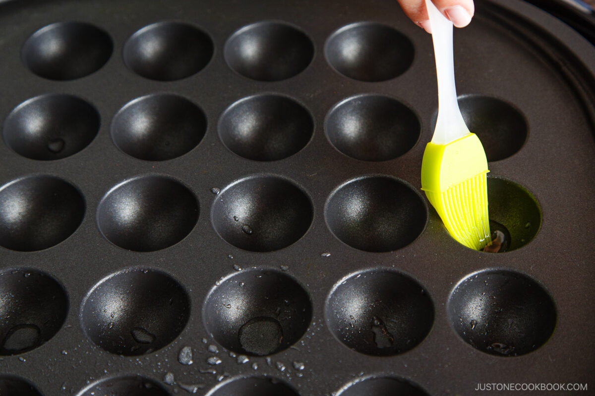 A hand uses a green silicone brush to oil the molds of a black takoyaki pan, preparing it for cooking.