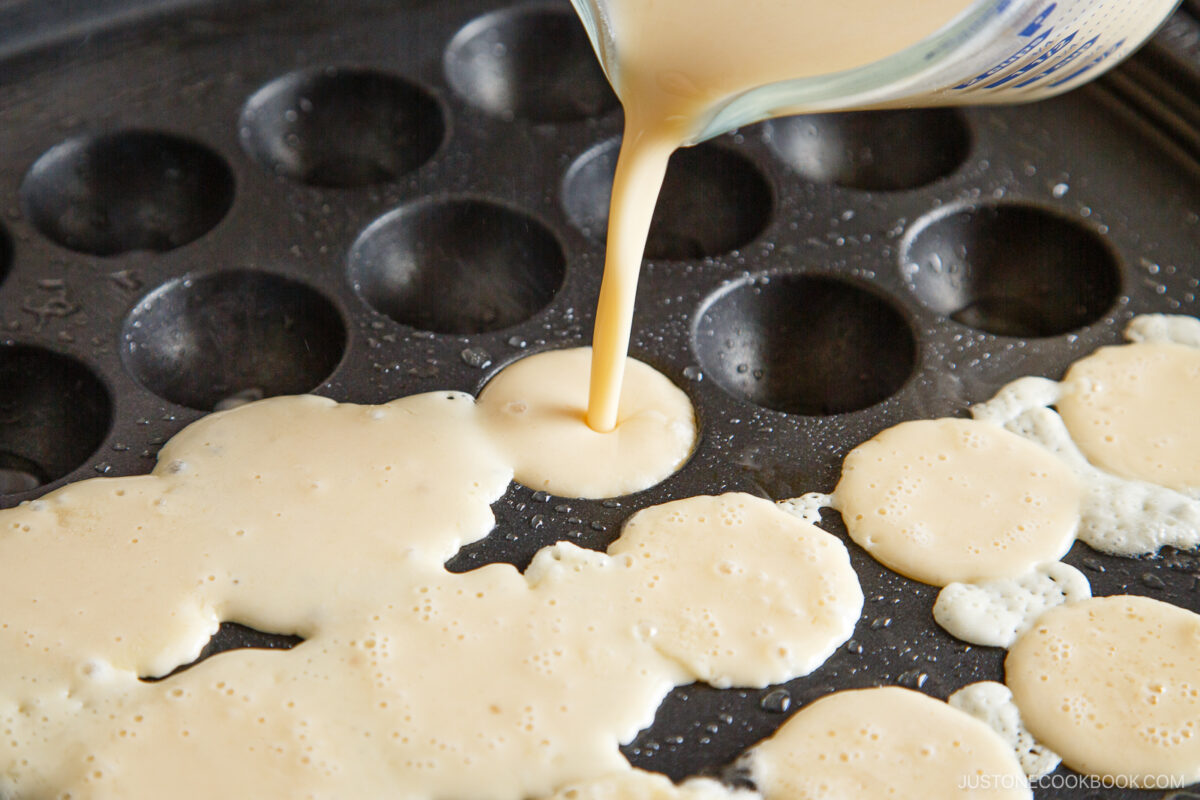 A close-up of pancake batter being poured from a measuring cup onto a takoyaki pan, filling one of the round molds while batter spreads over the cooking surface.