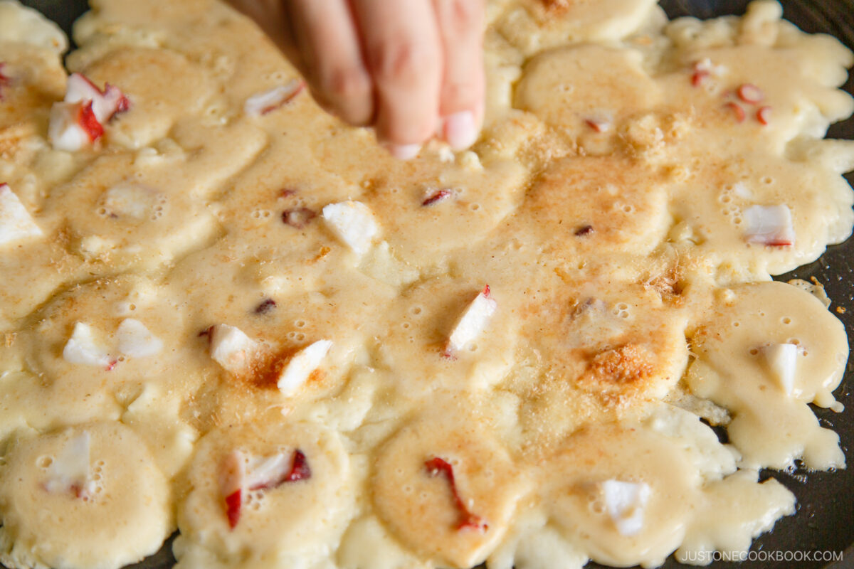 A hand sprinkles seasoning over a pan of uncooked batter with visible pieces of red and white octopus, preparing takoyaki.