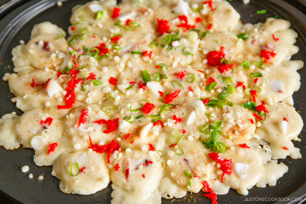 A close-up of takoyaki batter cooking on a griddle, topped with chopped octopus, green onions, red pickled ginger, and tempura bits. The mixture is bubbling and partially cooked, with colorful toppings scattered across.