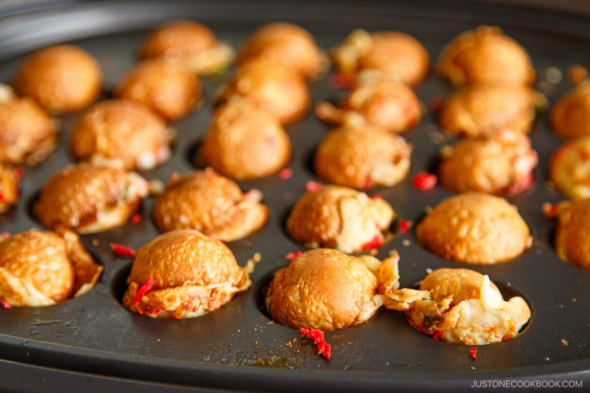 A close-up of golden-brown takoyaki balls cooking in a takoyaki pan. Some batter has seeped out around the balls, and bits of red filling are visible. The pan is partially filled, with some empty circular molds.