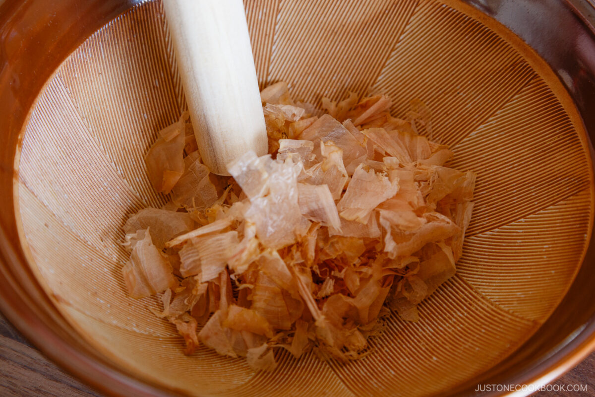 A close-up of a wooden pestle crushing light, thin bonito flakes in a ridged brown mortar.