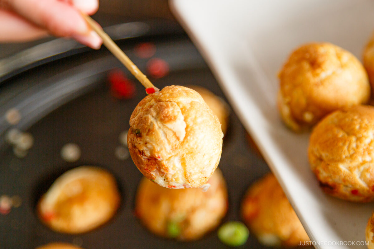 A hand holds a golden-brown takoyaki ball on a skewer above a plate with more takoyaki balls, with a takoyaki pan visible in the background.