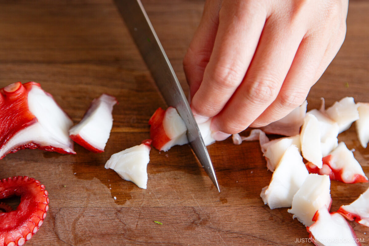 A hand uses a sharp knife to slice cooked octopus on a wooden cutting board, with pieces of red and white octopus scattered nearby.